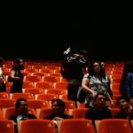 People seated in an empty theater with orange chairs.