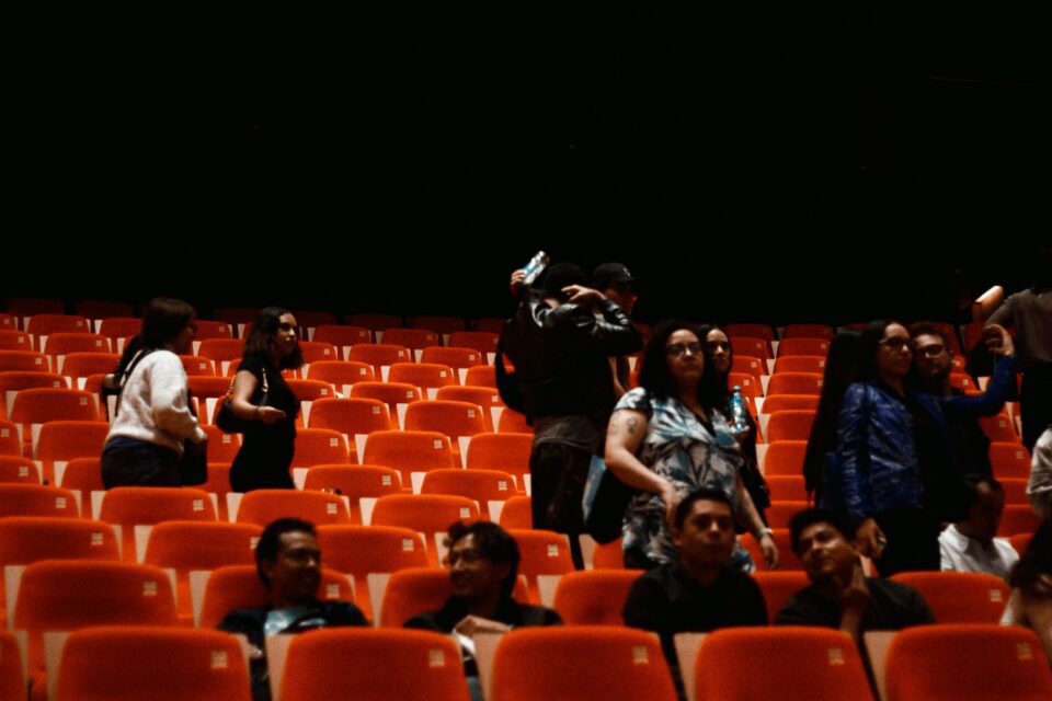 People seated in an empty theater with orange chairs.