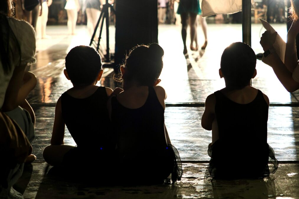Three children in black leotards sitting on a stage floor during a rehearsal